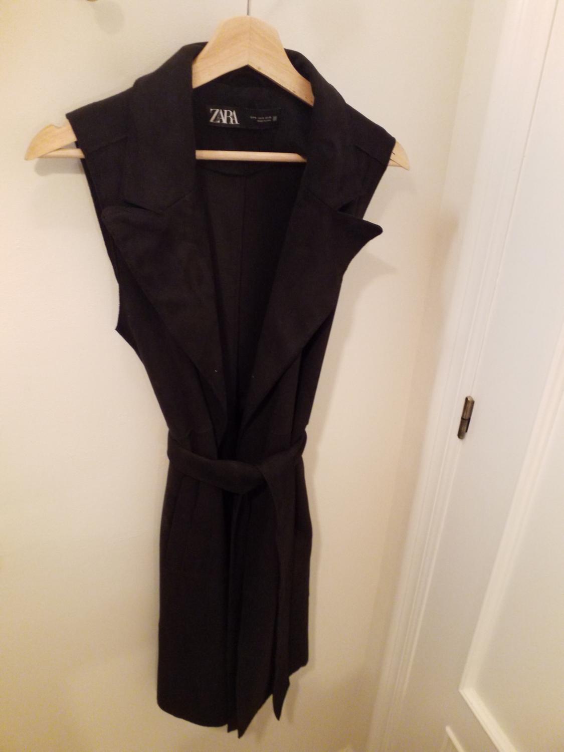 Black sleeveless waistcoat with belt, hanging on a wooden hanger. Visible label with the text "ZARA". White background.
