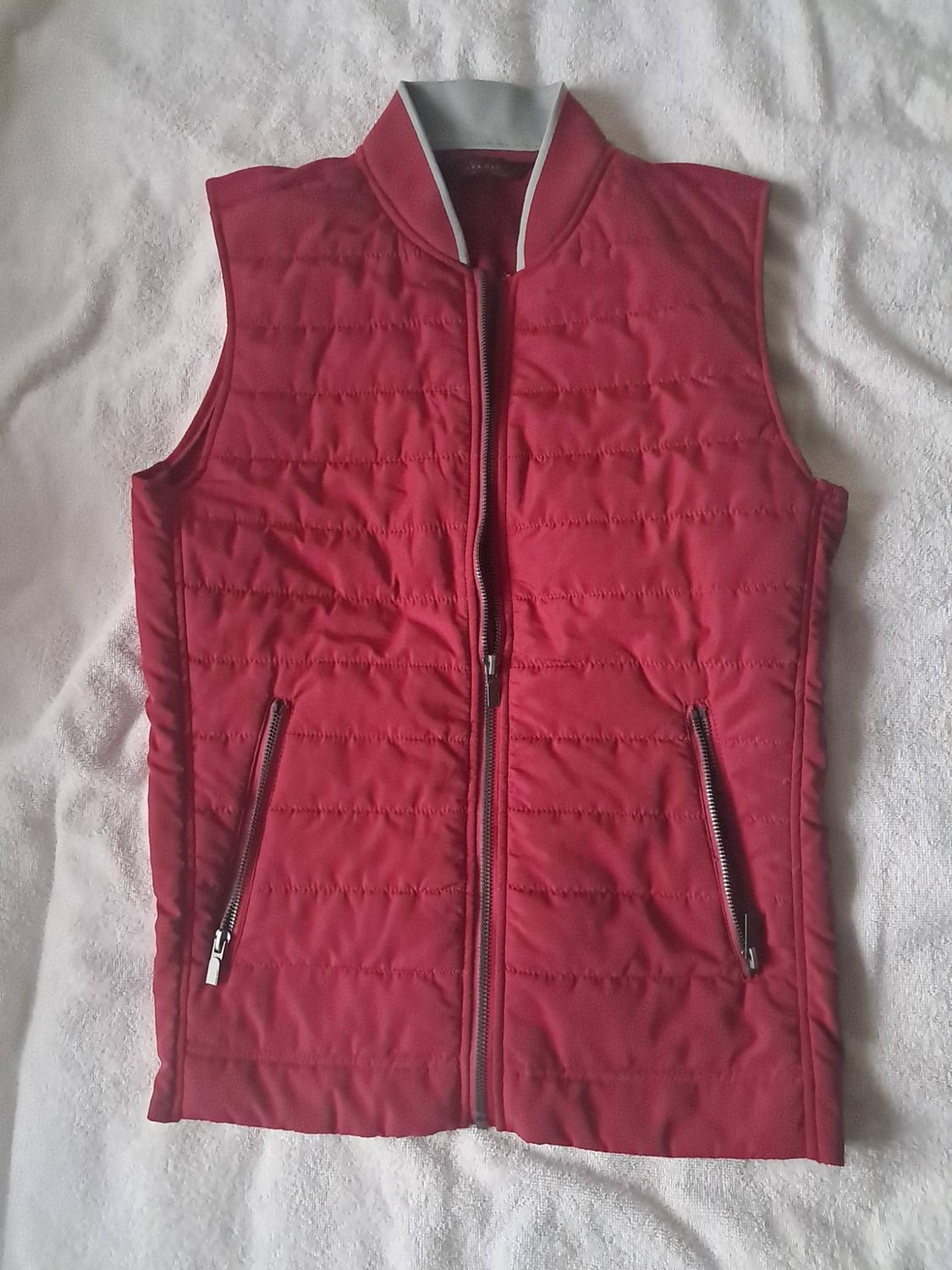 Red padded gilet with a grey collar, front zip fastening and zipped pockets, against a white background.