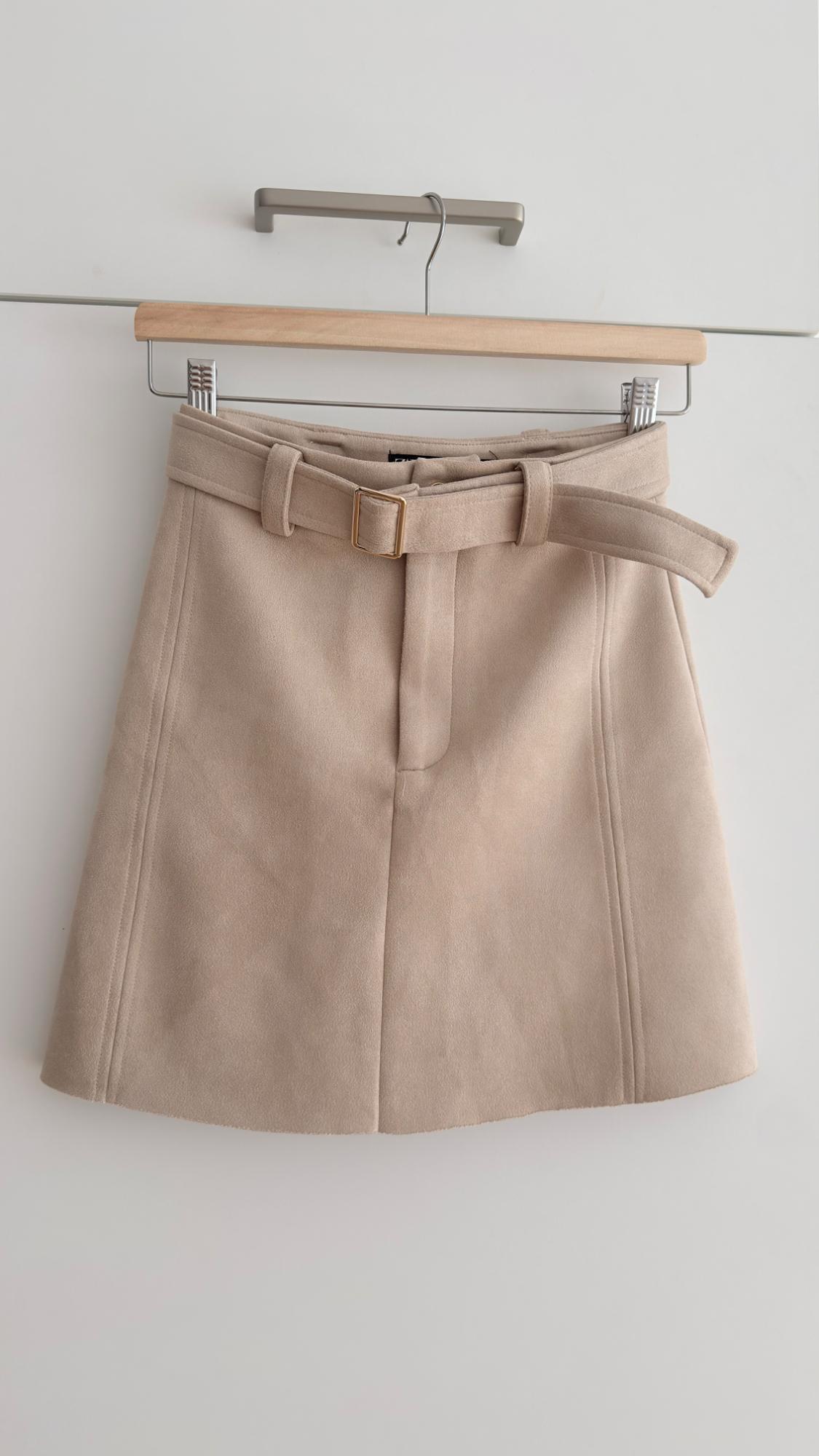 Short beige skirt with a belt, hanging on a wooden hanger against a white background.