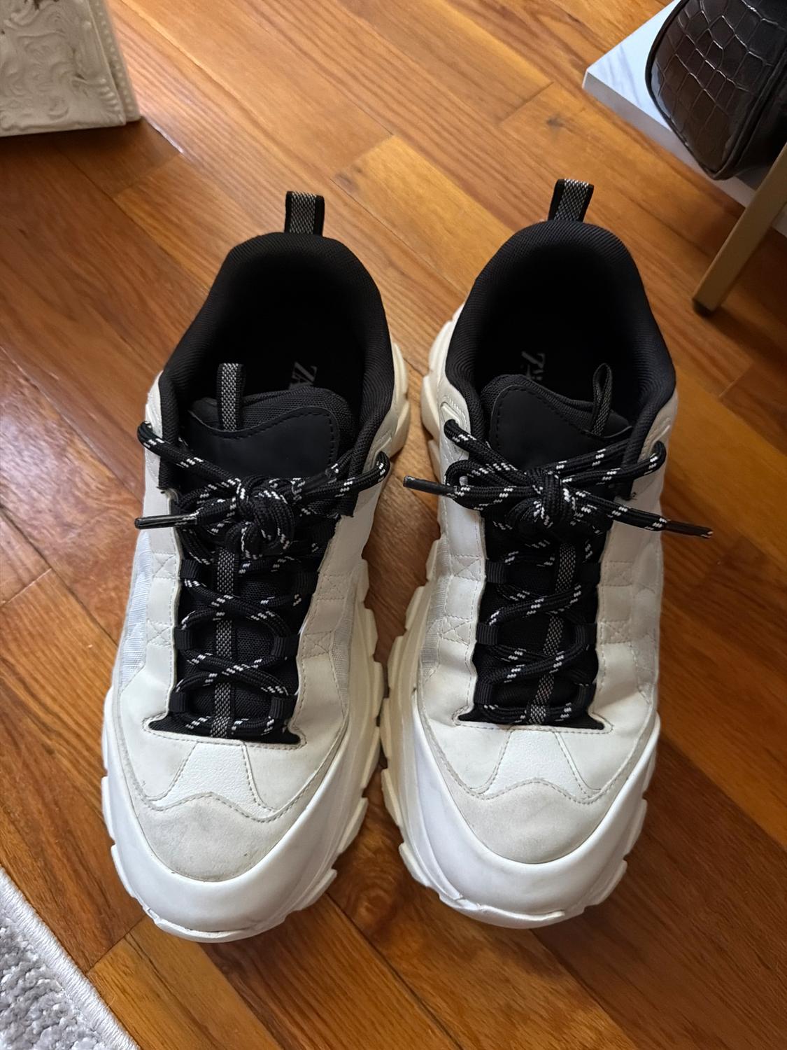 White athletic sneakers with a thick sole, black interior and laces, resting on a wooden floor.