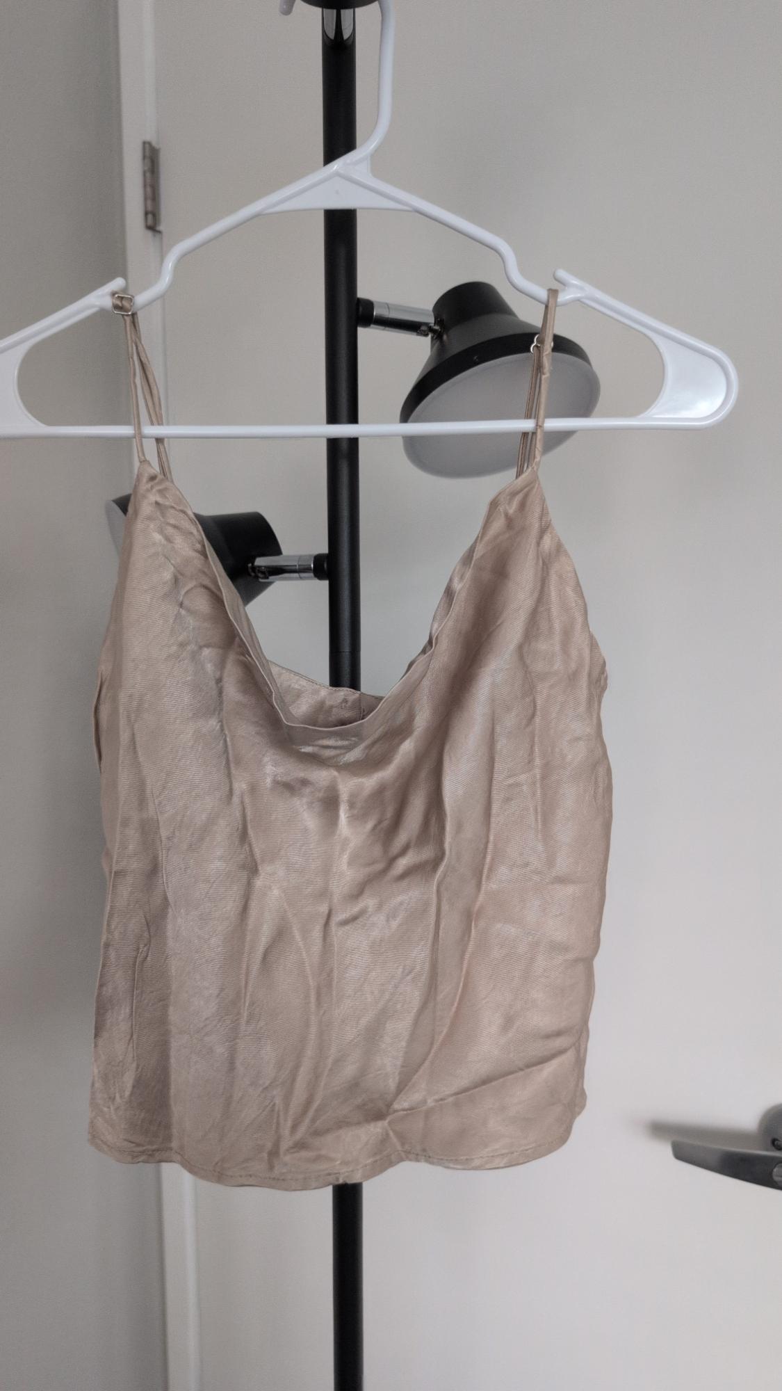 Beige satin camisole top with thin straps hanging on a white hanger over a black floor lamp, white wall background.