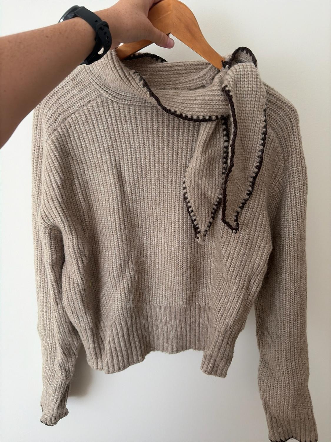Hand with watch holding a hanger with a chunky knit beige jumper. Wearing a scarf around the neck with brown stitched edges. White background.