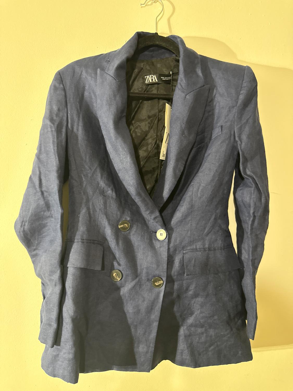 Double-breasted blue linen jacket, hanging on a hanger against a yellow background. Visible label with the ZARA brand.