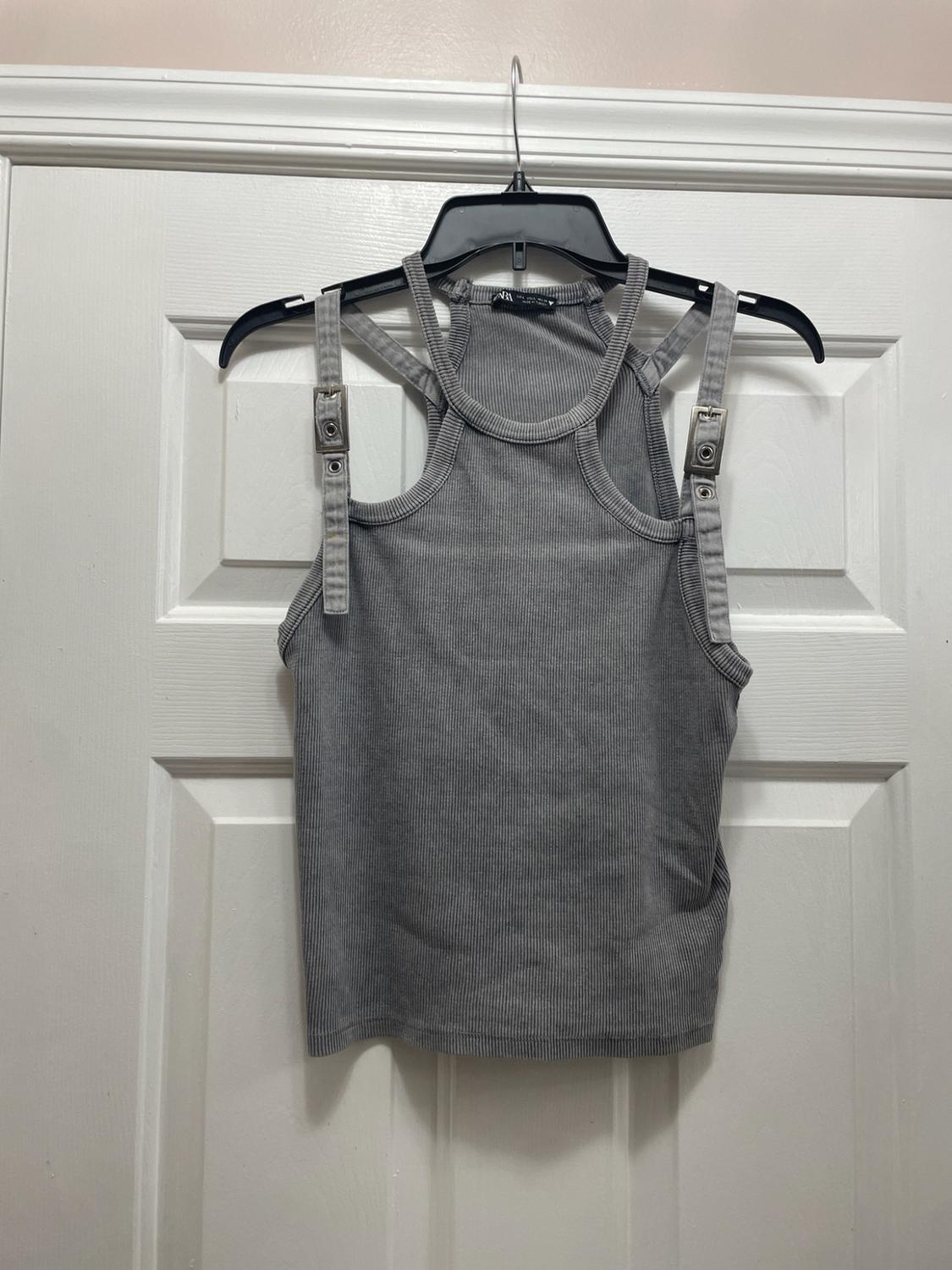Gray ribbed tank top with double straps and metallic details, hanging on a black hanger in front of a white door.