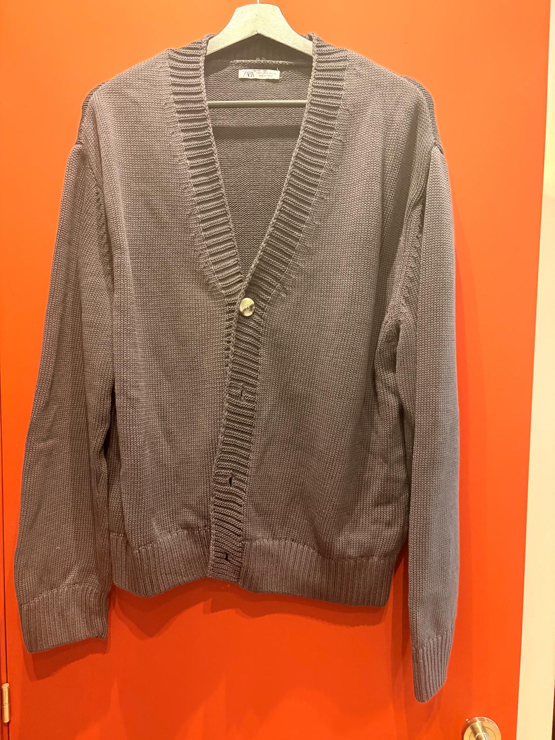 Grey knitted cardigan hanging on a hanger over an orange door. Label visible on the collar.