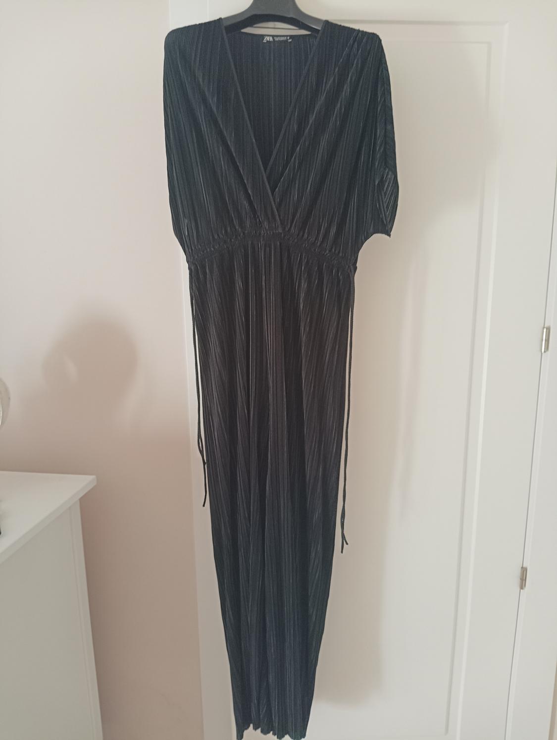 Black pleated dress hanging on a hanger over a white door. The label reads "Zara Woman".
