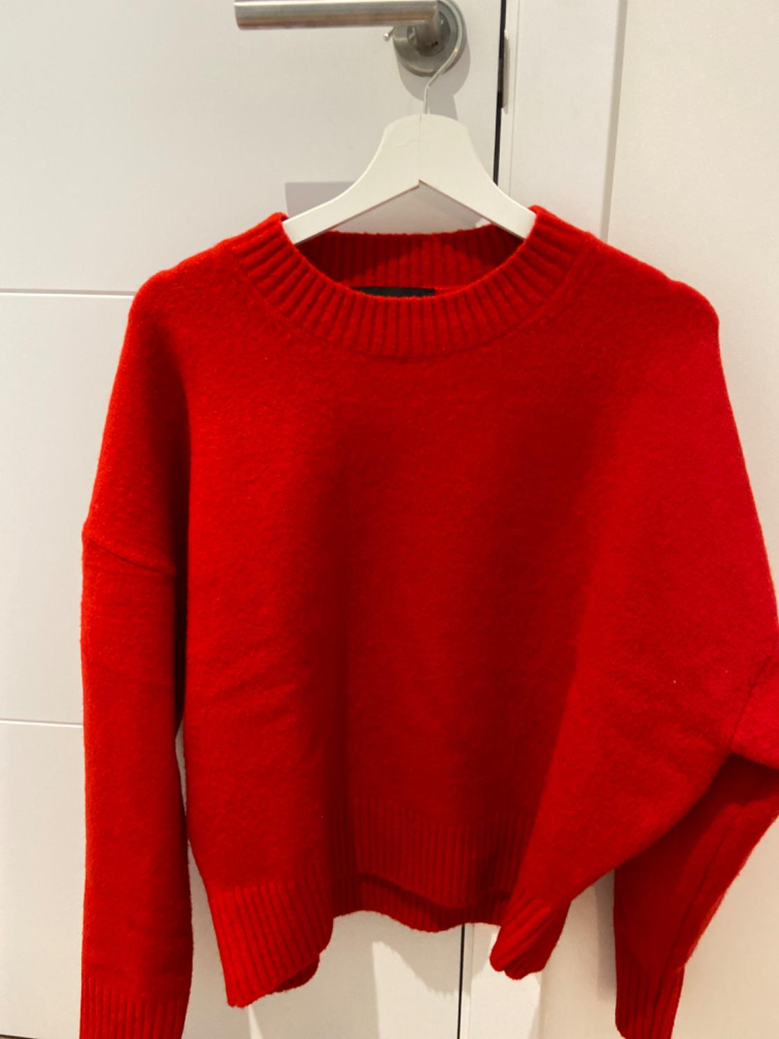 Chunky knit red jumper hanging on a white hanger over a white door with a silver handle.