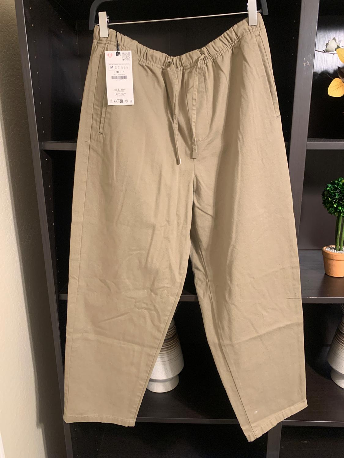 Beige trousers with elasticated waist and drawstring, hanging on a dark shelf. Visible tag with size M and price in dollars.
