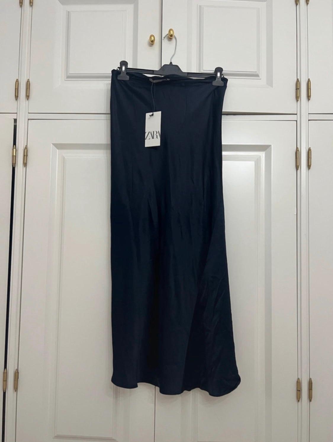 Dark satin skirt on a hanger hanging from the handle of a white wardrobe. Label with the text ZARA. Relaxed home atmosphere.