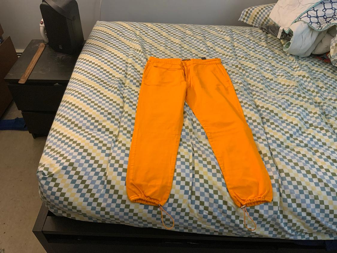 Orange pants with ankle drawstrings on a bed with diamond-patterned sheets. Casual bedroom setting with a nightstand.