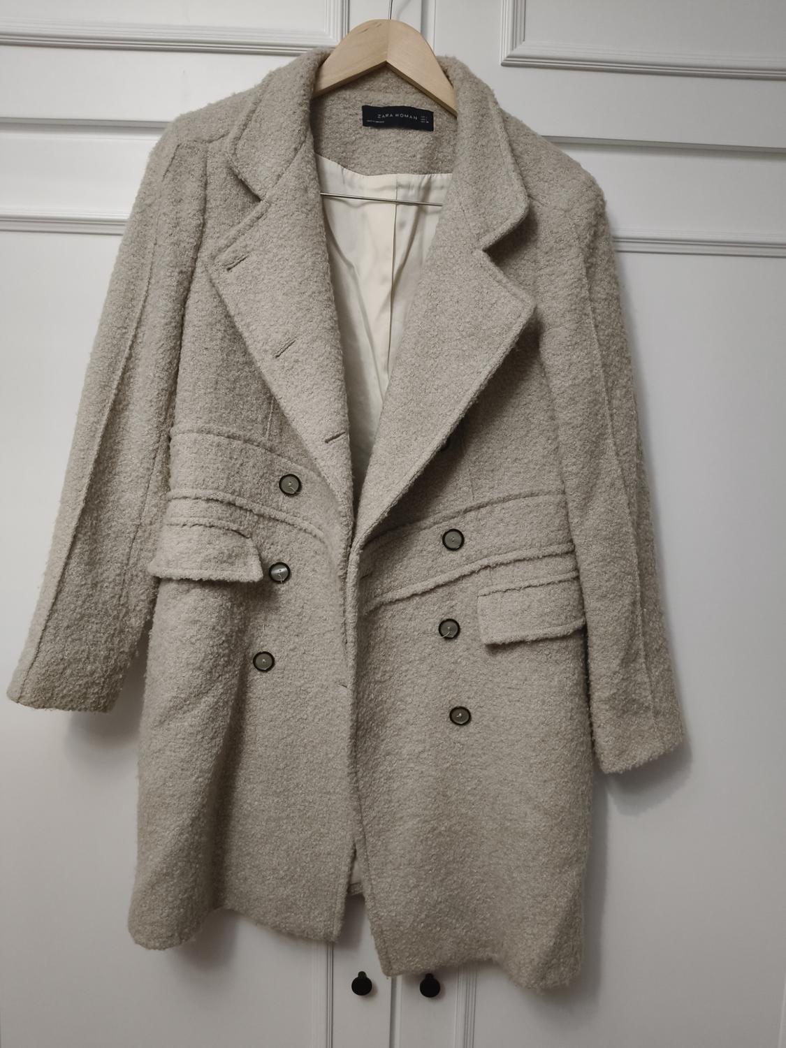 Beige wool coat hanging on a wooden hanger, with dark buttons and wide lapels, against a background of white doors.