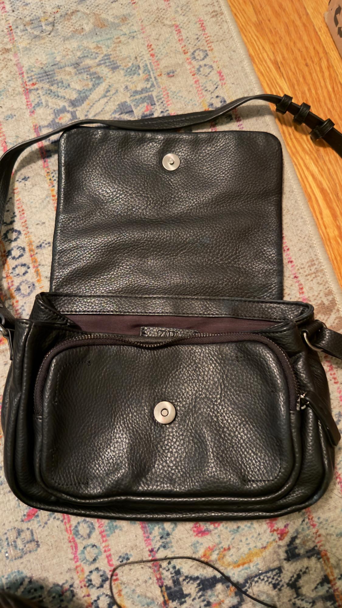 Black leather handbag with an open flap and a metallic button closure on a colourful rug. Visible adjustable strap.