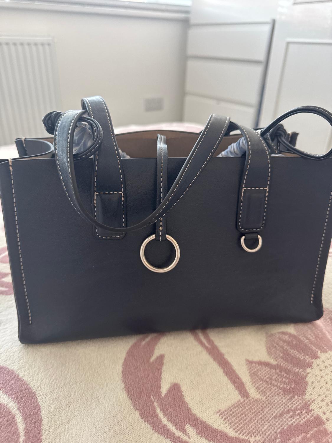 Black handbag with white stitching and metallic rings, on a floral-patterned surface in shades of pink. Room background.