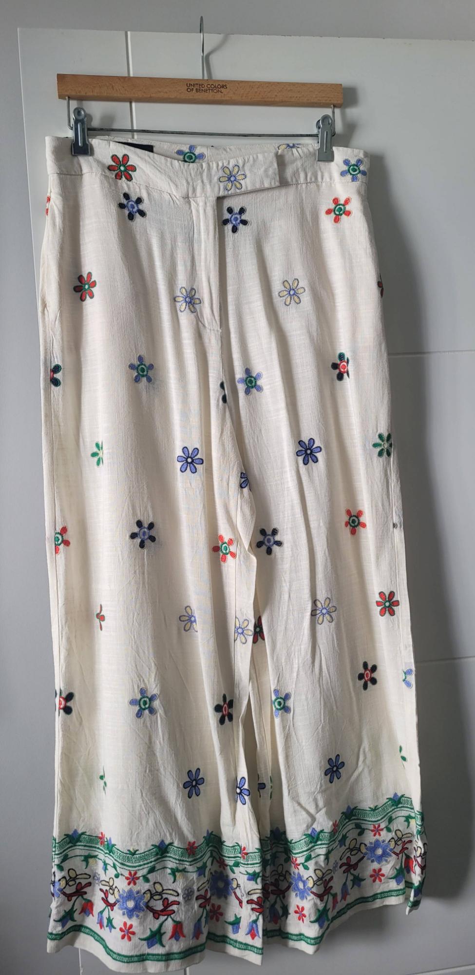 White trousers with multicoloured floral embroidery, hanging on a wooden hanger with the text "United Colors of Benetton".