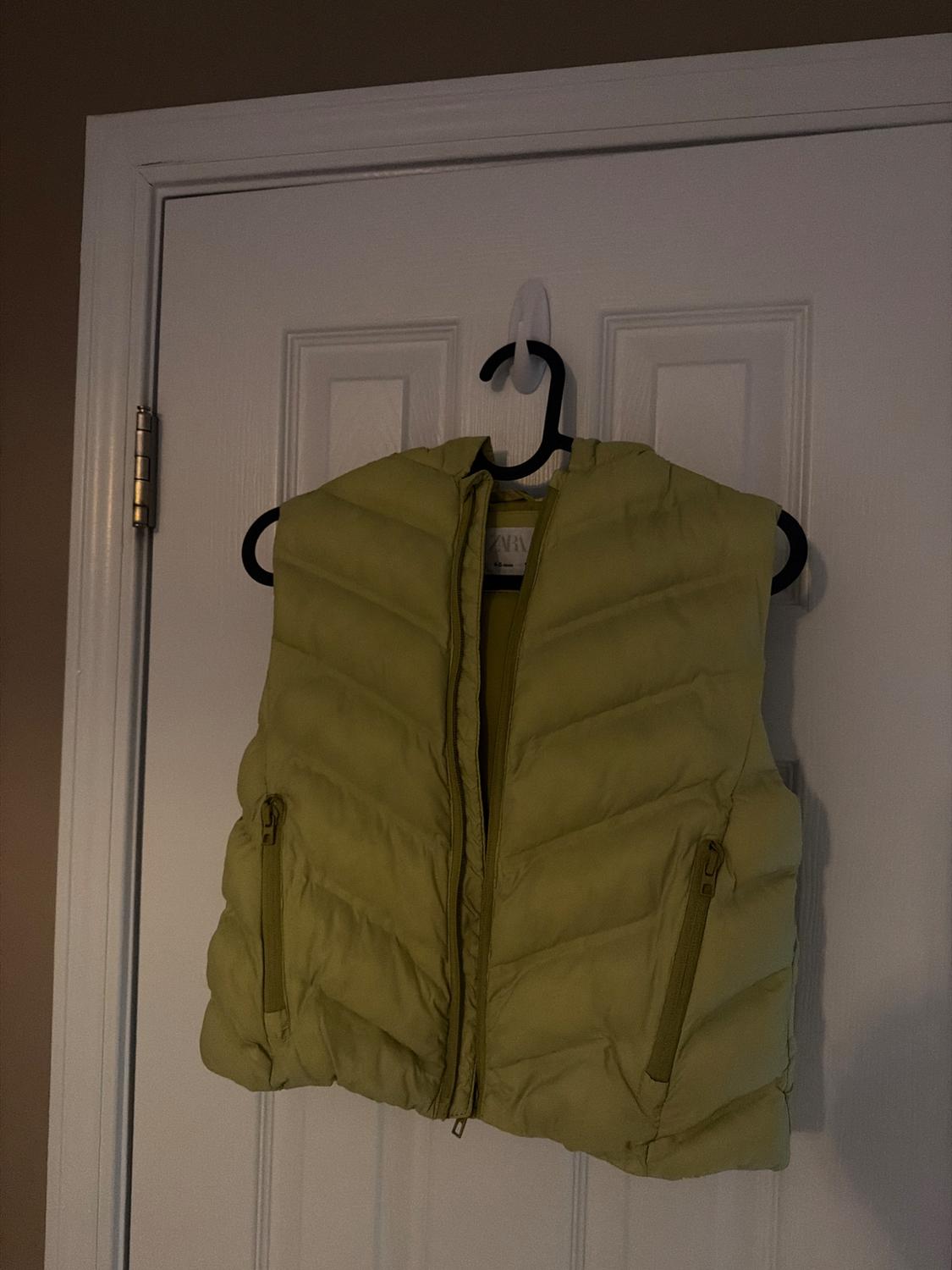 Light green padded vest hanging on a white door, with front closure and zip pockets.