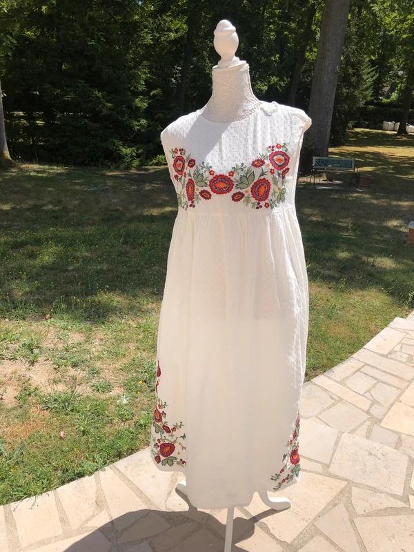 Μανεκέν σε εξωτερικό χώρο με λευκό αμάνικο φόρεμα και κόκκινα και πράσινα λουλουδένια κεντήματα στο στήθος και στο τελείωμα, σε πέτρινο αίθριο με κήπο στο βάθος.