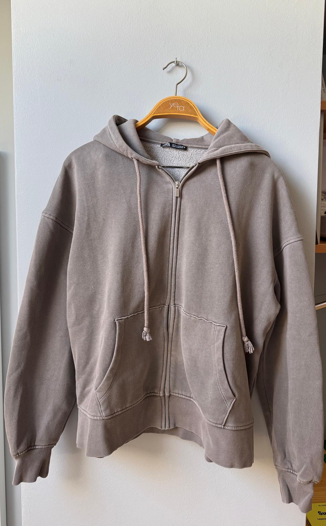 Light grey hoodie hanging on a wooden hanger with the "yera" brand. White background and plain wall.
