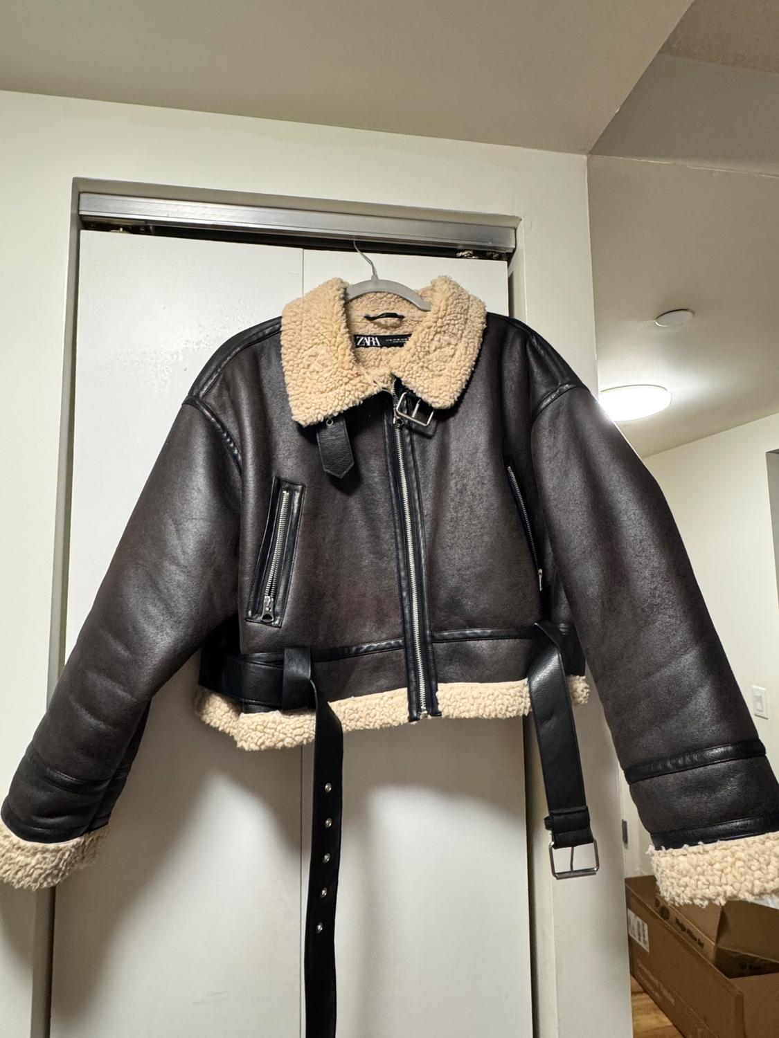 A brown leather jacket with a beige wool interior, belt, and zippers is hanging on a white door in a well-lit space.