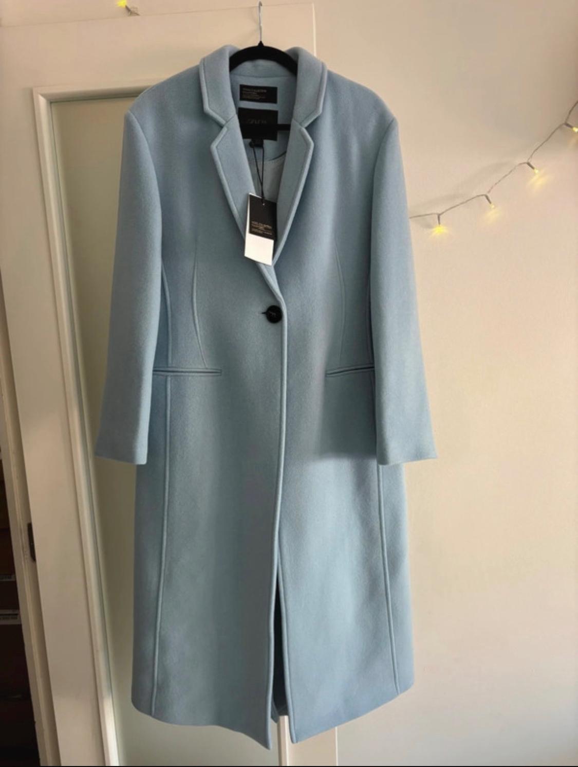 Light blue long coat with tags, hanging on a white door. It has lapels and a single black button. Background features a warm fairy light garland.