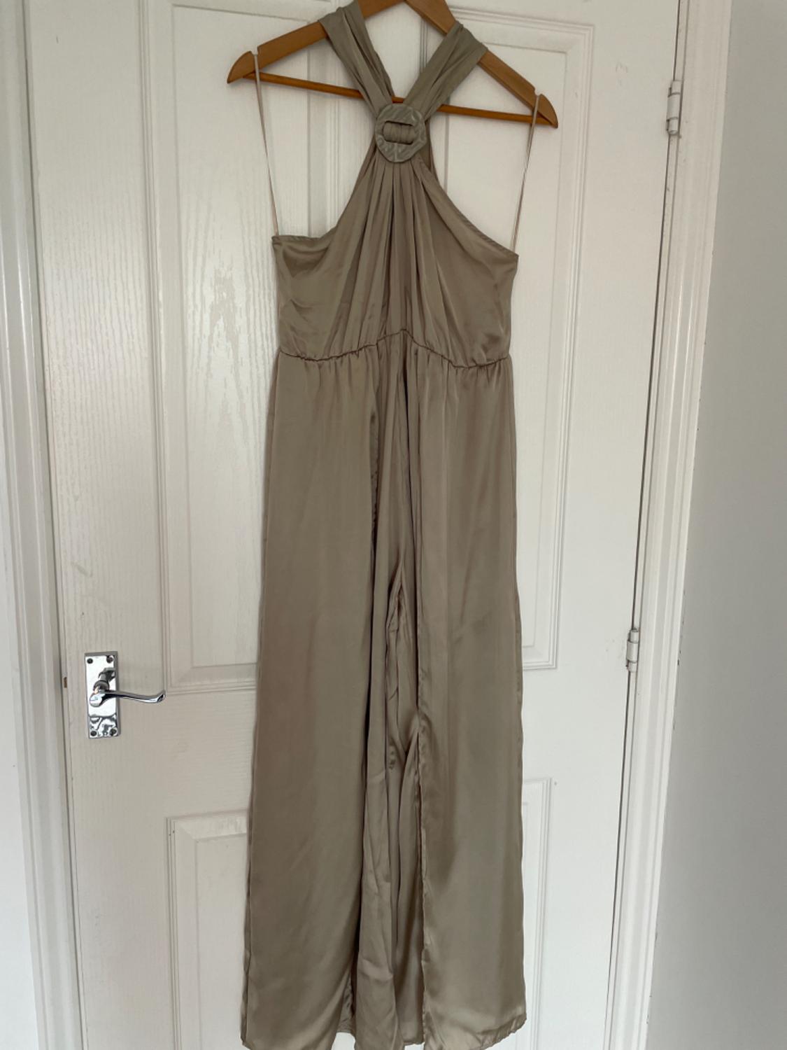 Beige satin long jumpsuit with a halter neck and circular detail, hanging on a wooden hanger against a white door. Relaxed style.