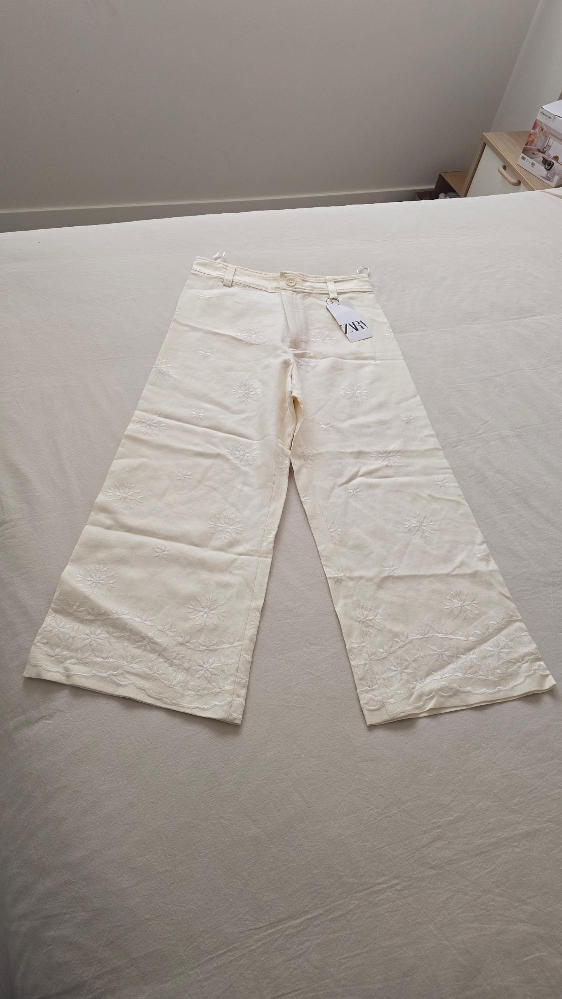 White wide-leg trousers with floral embroidery, visible ZARA logo tag, on a beige bed in a minimalist room.