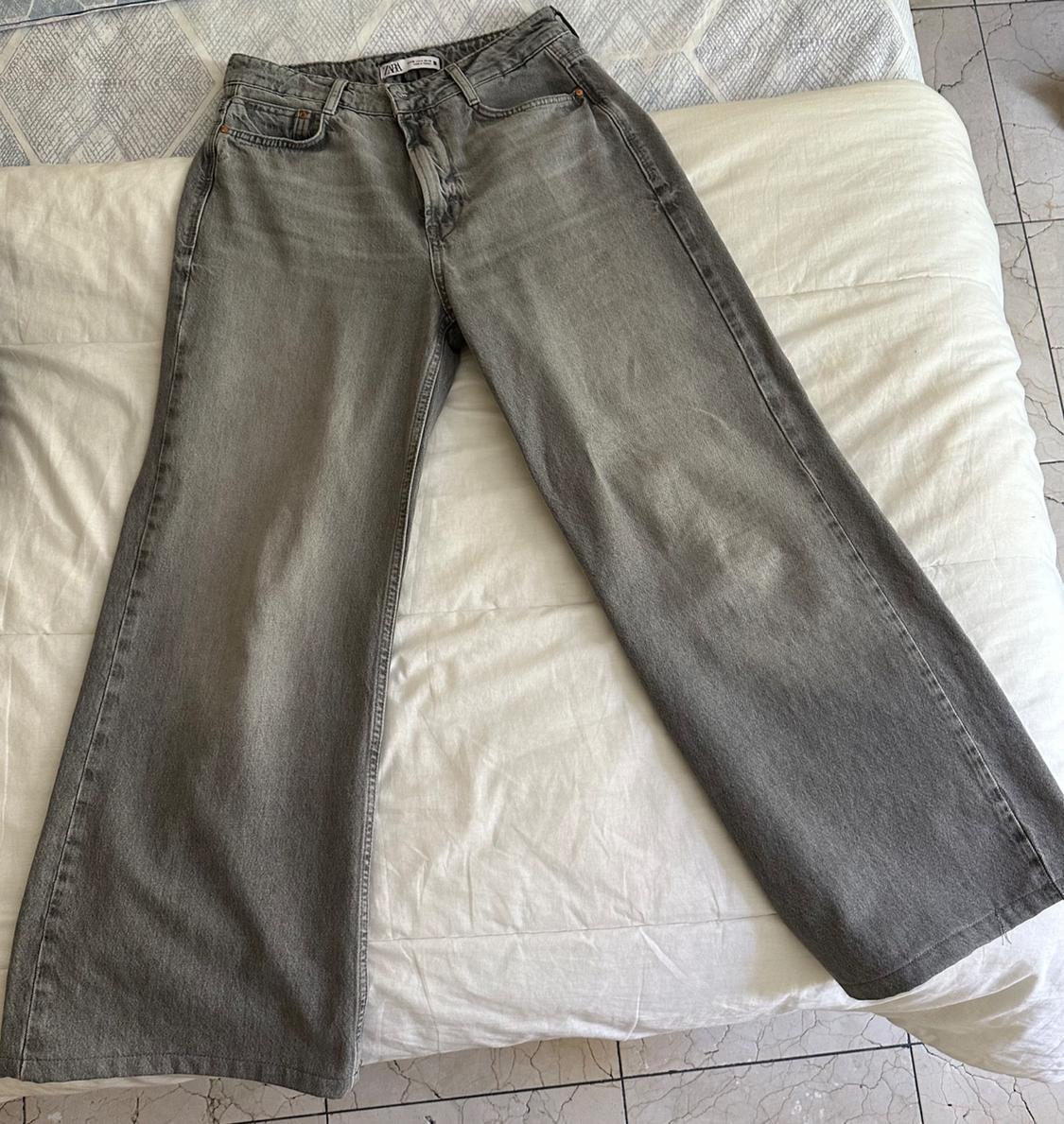 Grey wide-leg denim trousers on a white bed, with visible label on the waistband.
