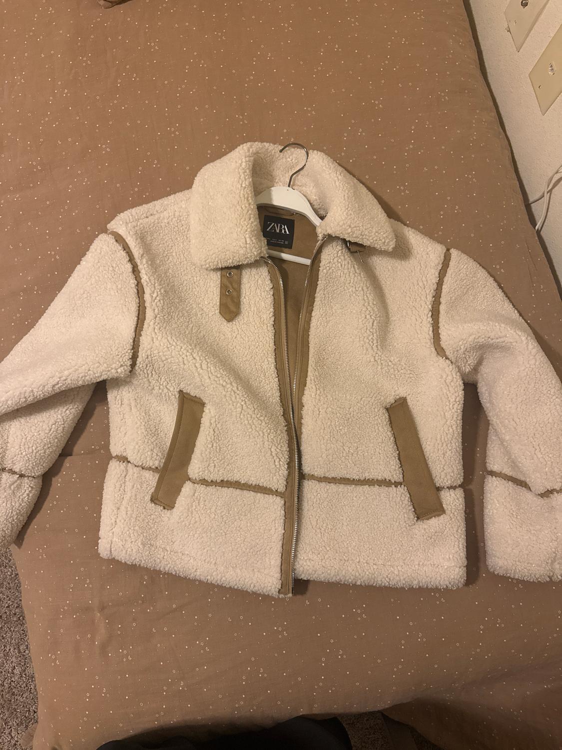 White wool jacket with brown detailing, front zip, and high collar, hanging on a hanger over a beige bed.
