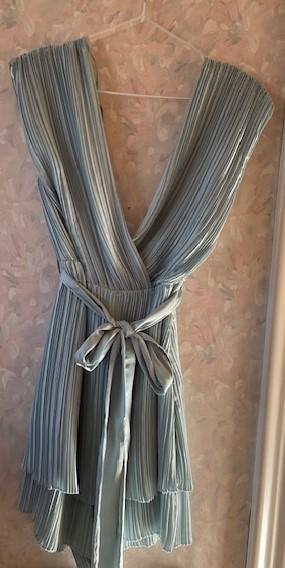 Light blue pleated dress hanging on a hanger against a floral wallpaper background. It has a knotted belt at the waist.