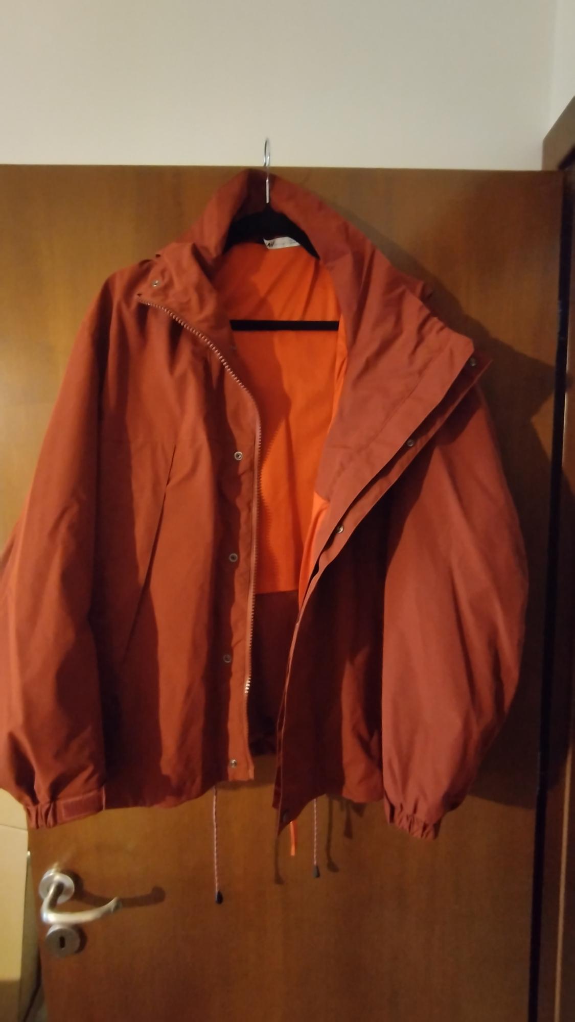 Red jacket hanging on a wooden door, with visible orange lining. The jacket has a hood and adjustable drawstrings.