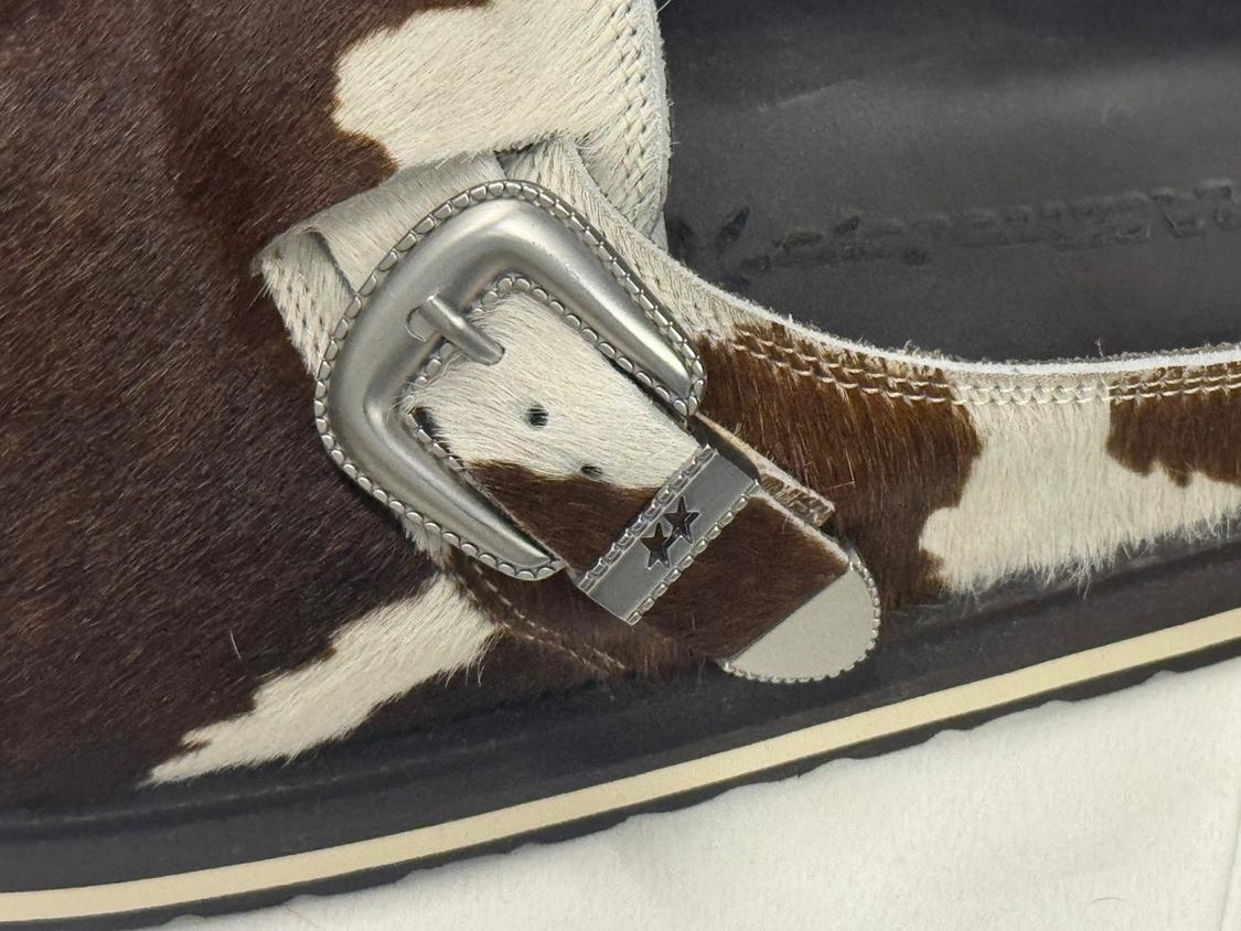 Close-up of a sandal with a pattern of brown and white spots, featuring a silver metal buckle decorated with two engraved stars.