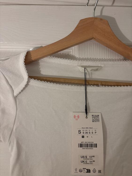 White t-shirt on wooden hanger with hanging price tag. Visible text: "US $17.90, UK £11.99, S 26 SSP".