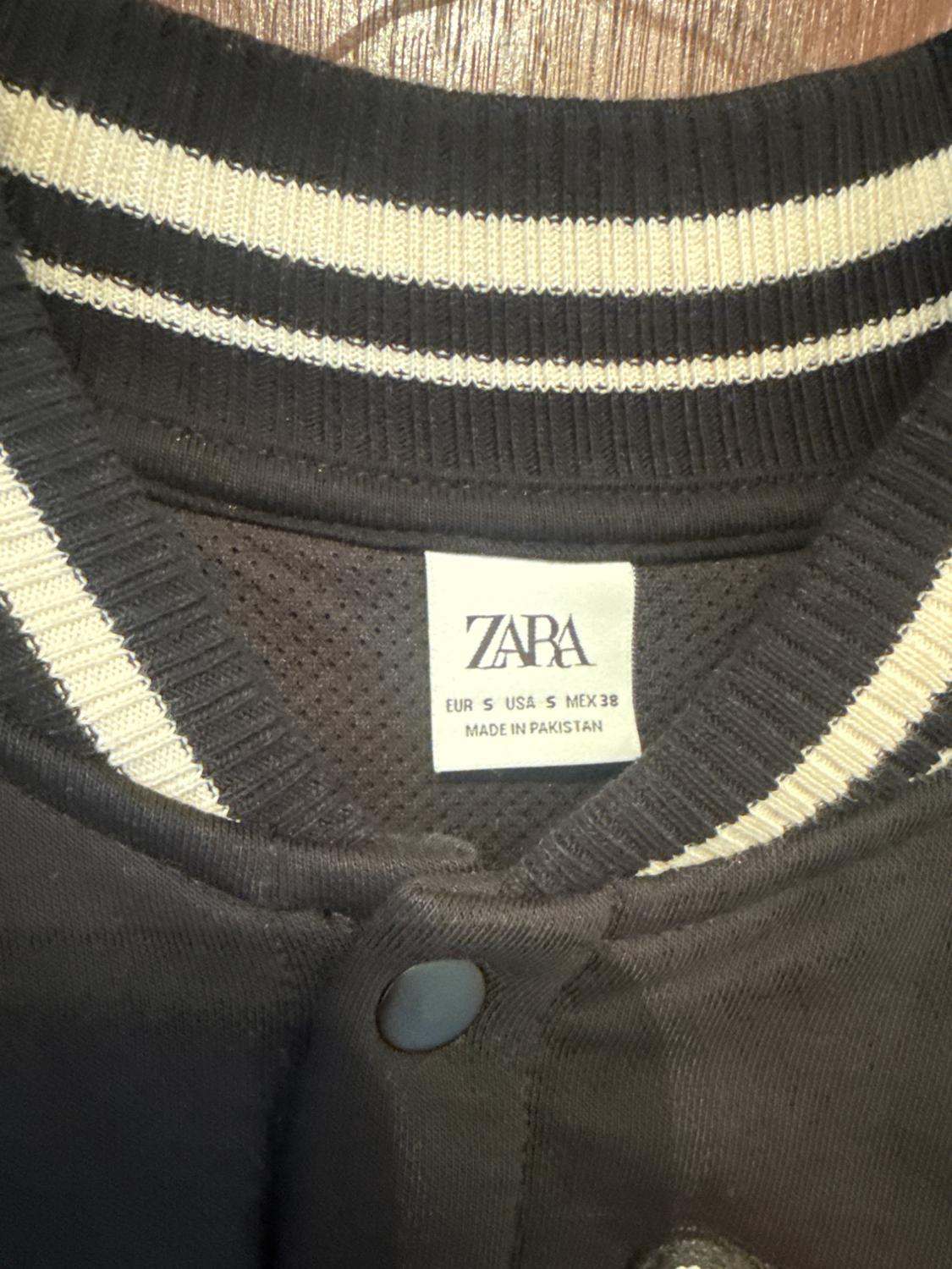 Black jacket with a collar featuring white stripes and a button. Label with text: ZARA EUR S USA S MEX 38 MADE IN PAKISTAN.