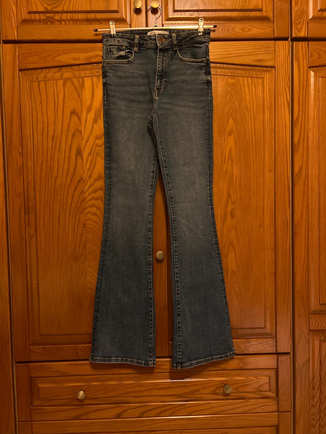 Blue flared jeans with a ZARA label, hanging on a clothes hanger in front of a wooden wardrobe.