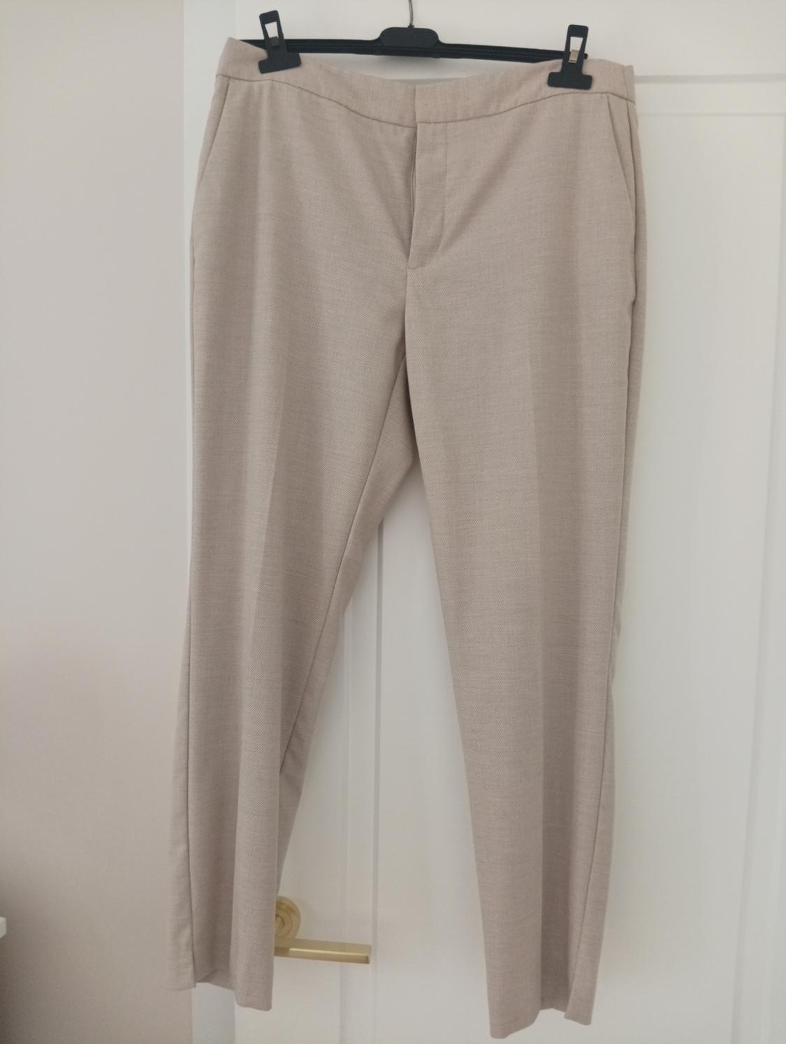 Beige trousers hanging on a black hanger over a white door. Plain fabric and straight cut, with side pockets.