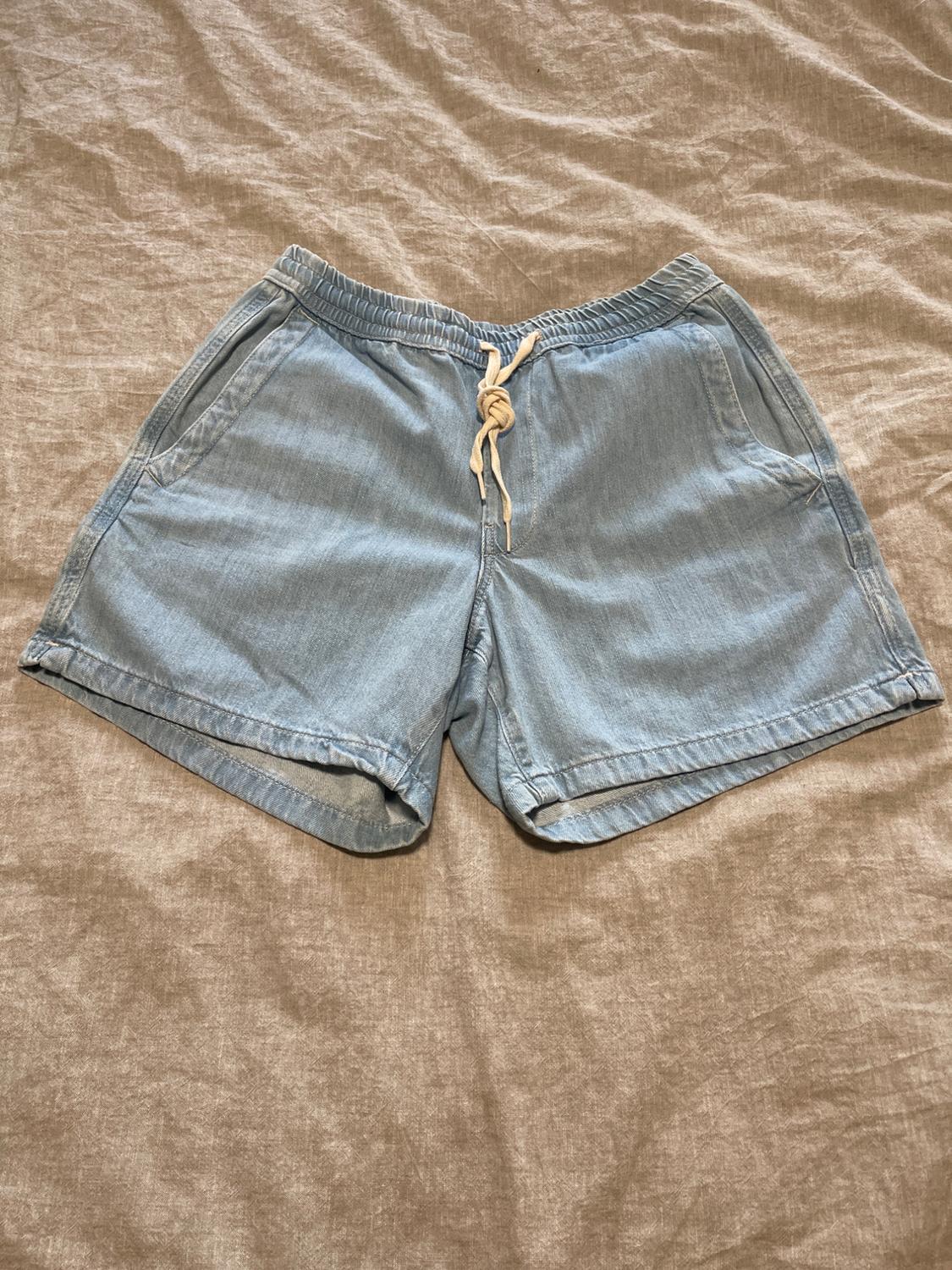 Light blue denim shorts with an elasticated waist and beige drawstring, on a wrinkled beige fabric.