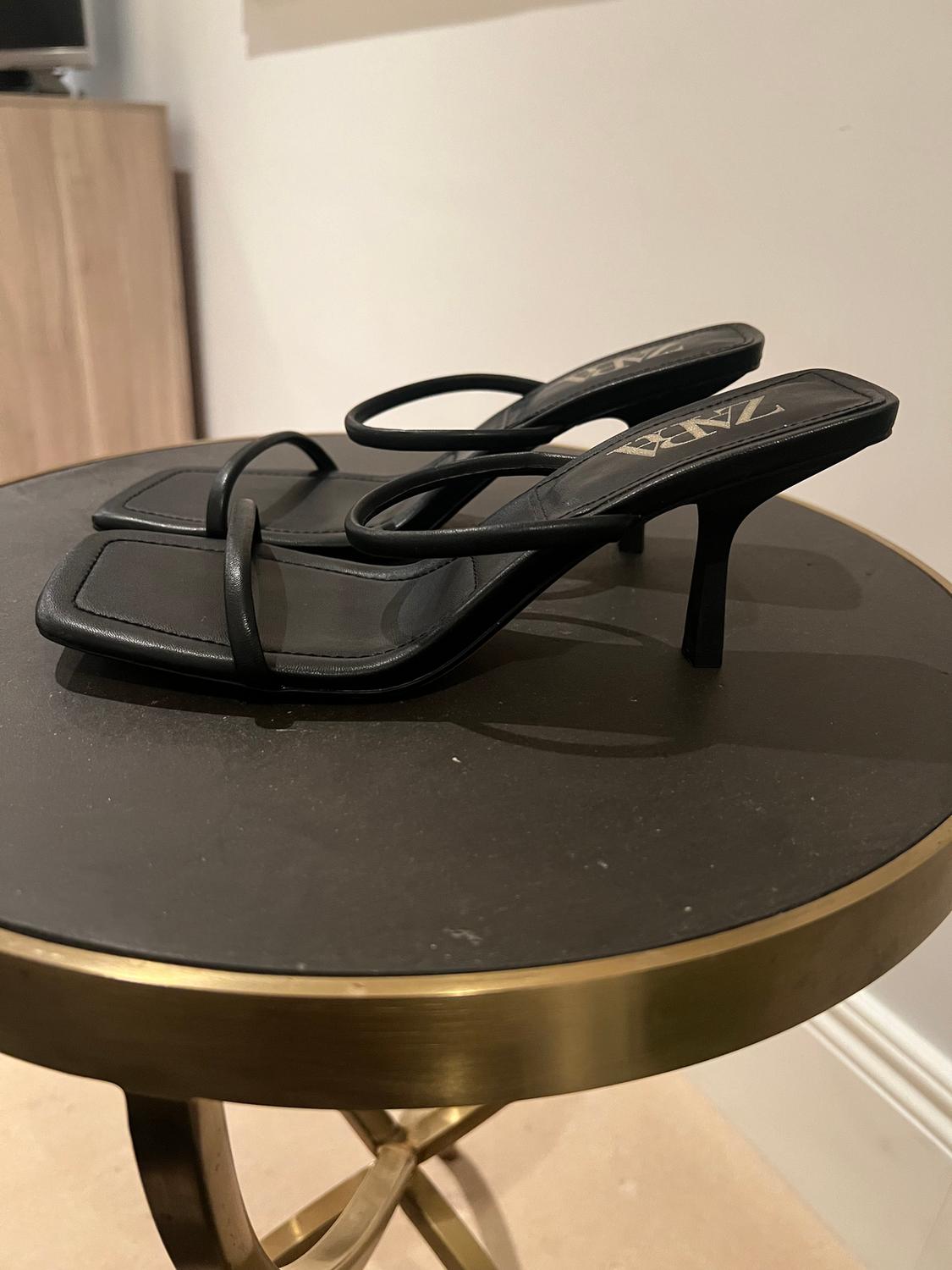 Black low-heeled sandals with thin straps, visible branding on the insole, on a round gold metal table.