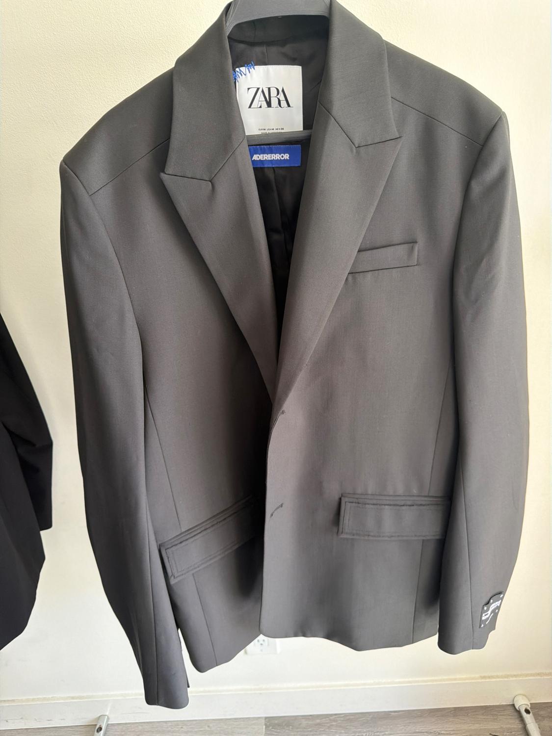 Grey jacket hanging on a hanger, with visible Zara and Adererror tags inside. White background and wooden floor.