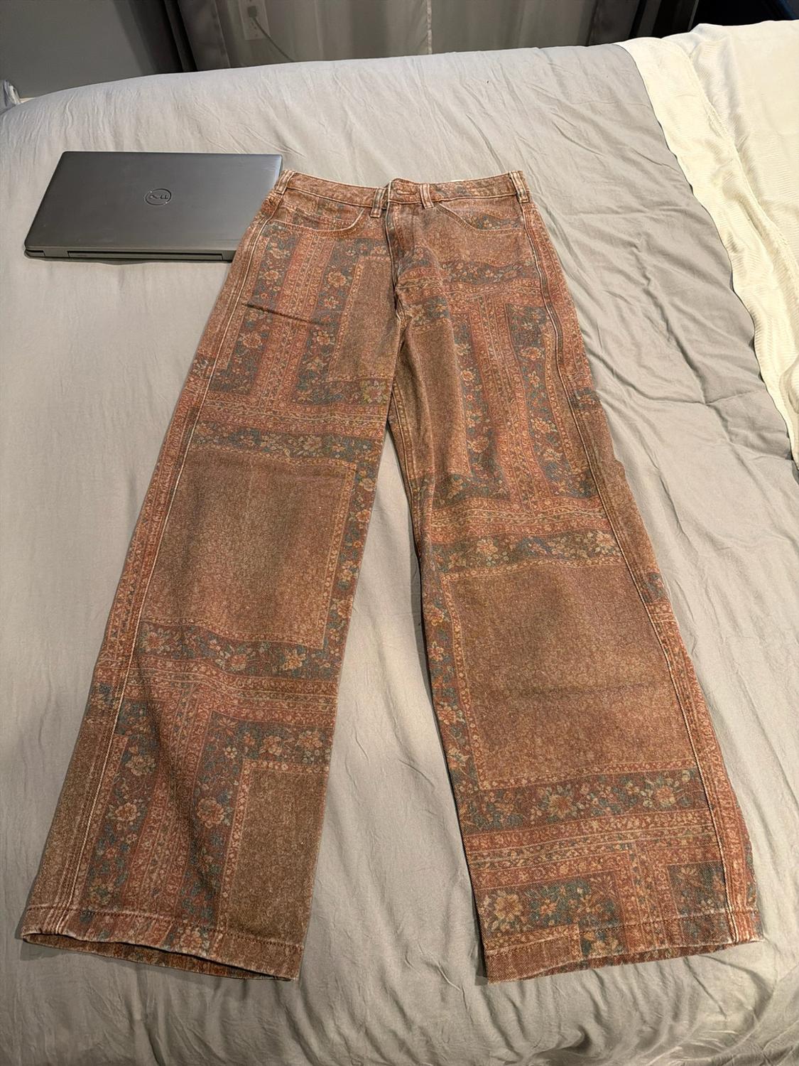 Wide-leg trousers with a tapestry-style print in brown tones, on a grey bed next to a closed laptop. Relaxed atmosphere.