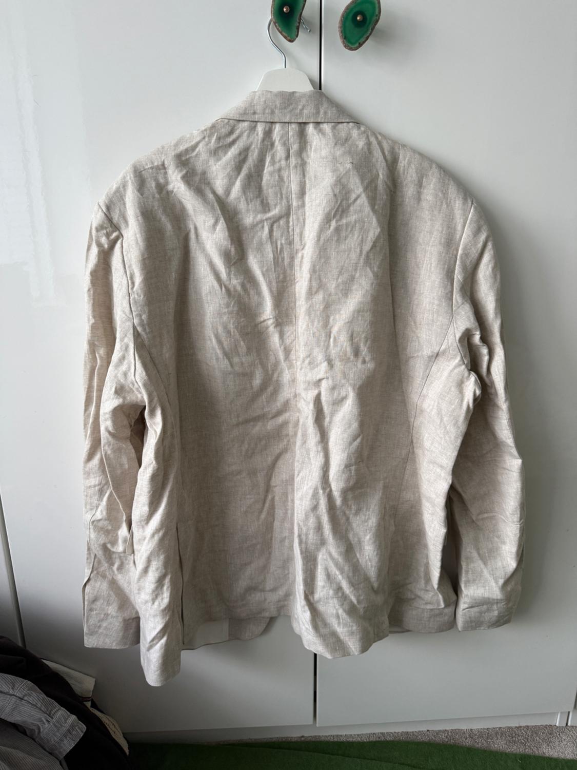 Creased beige linen jacket hanging on a white hanger in front of a white wardrobe. Bottom of clothing visible.