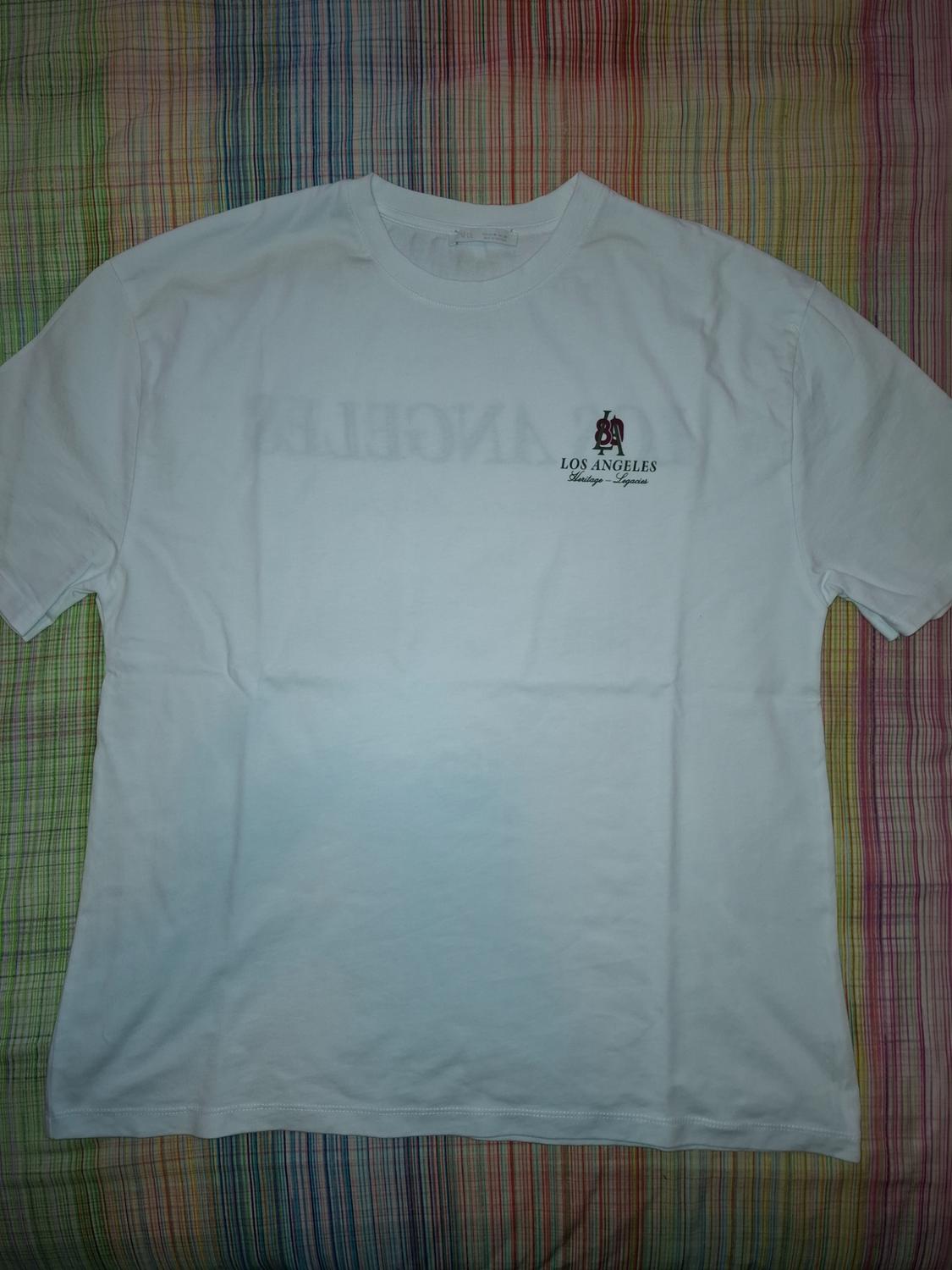 White T-shirt with "Los Angeles" text and logo on the chest, over a multicoloured striped fabric background.