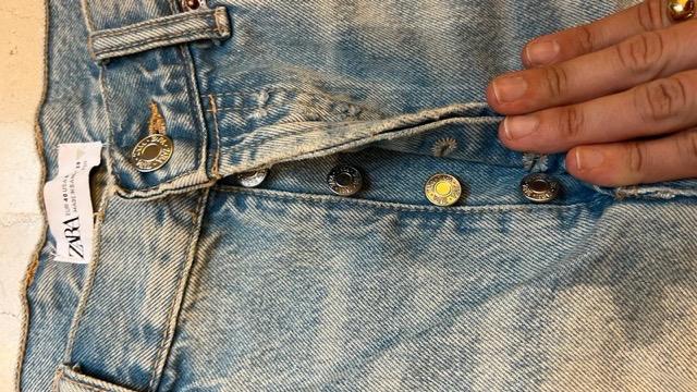 Hand displaying buttons of distressed blue jeans with visible label that reads "ZARA". Light background and denim texture.