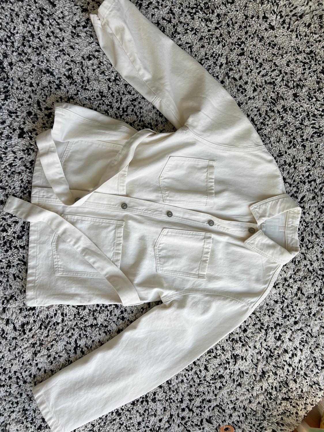 White, long-sleeved jacket with front pockets and buttons, placed on a thick, grey textured rug.