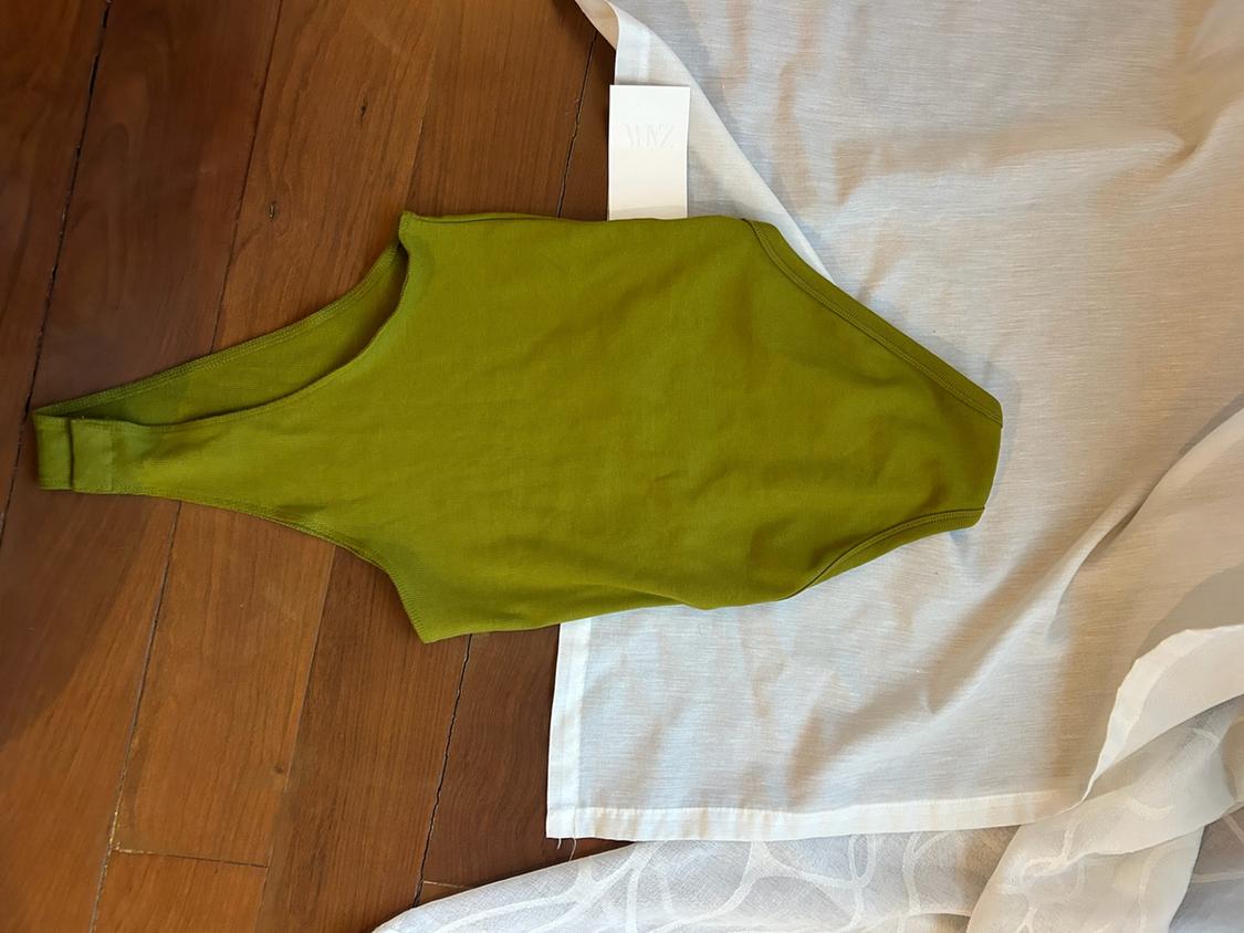 Green bodysuit with a ZARA label on a wooden floor next to a semi-transparent white fabric.