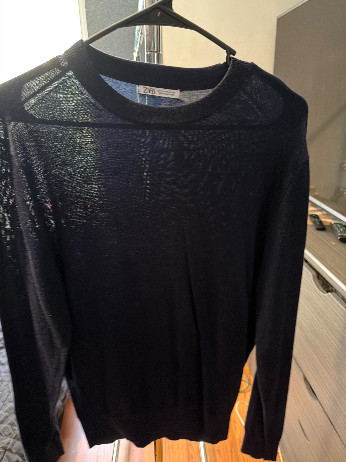 Dark long-sleeved sweater hanging on a hanger. Tag with ZARA text.
Room background with furniture and wooden floor.