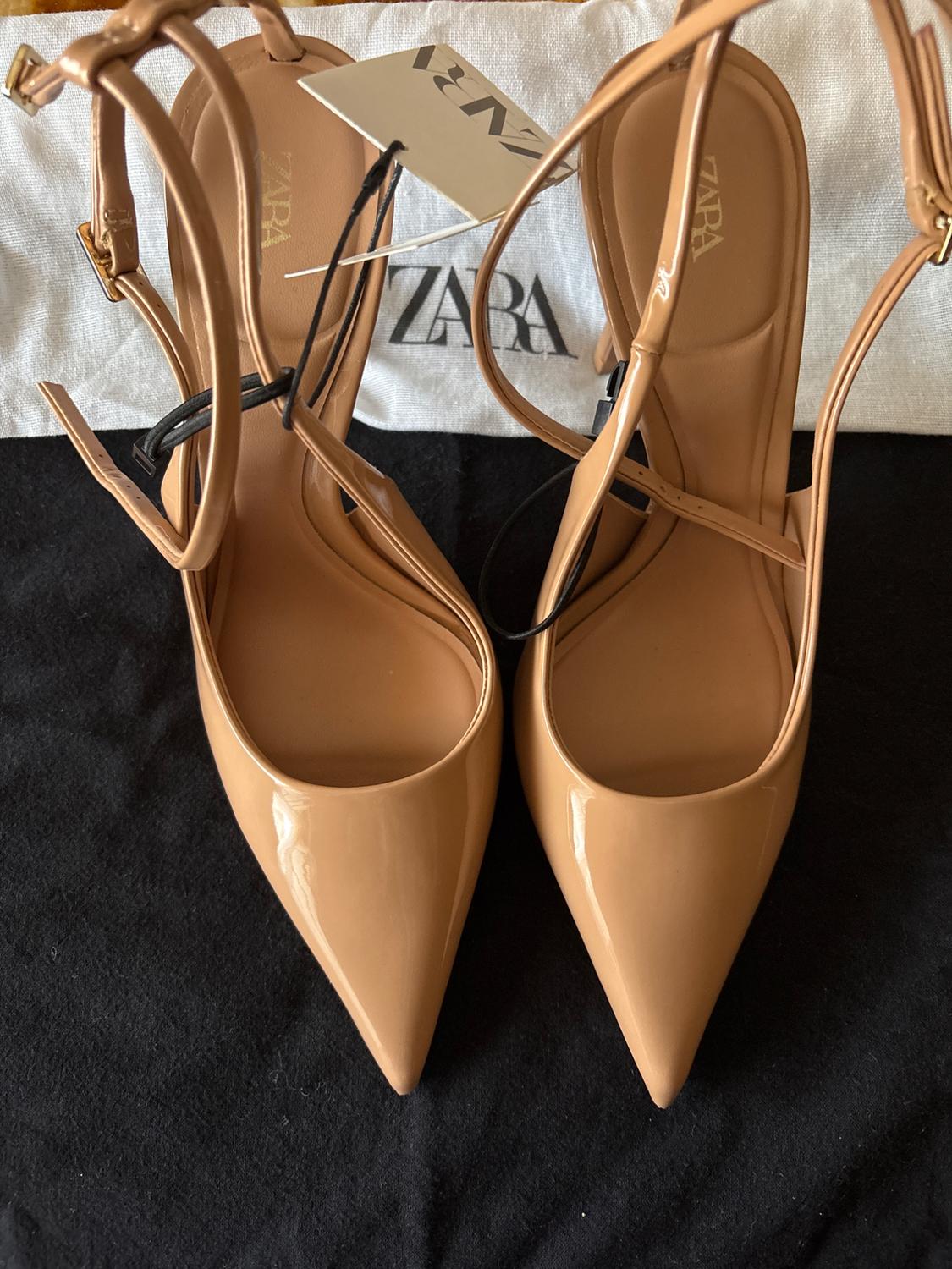 Beige high heels with pointed toes and straps, on black fabric. Label and fabric bag with "ZARA" logo visible.