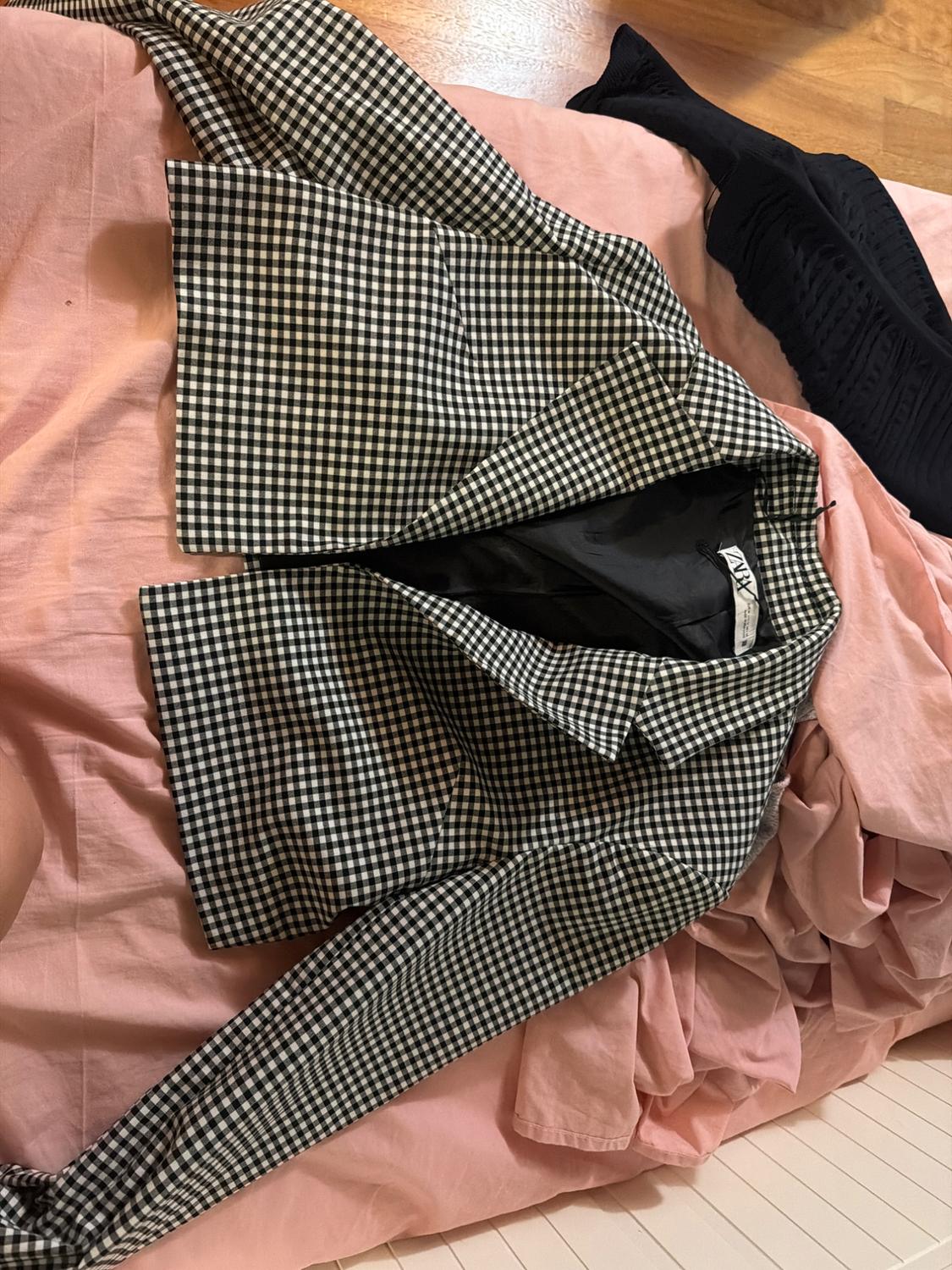 Black and white checked jacket with a ZARA label, on a crumpled pink sheet next to a dark garment. Relaxed vibe.