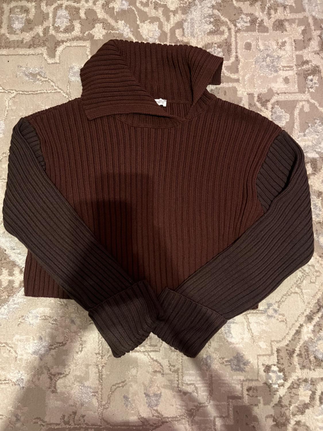 Dark brown knitted sweater with a high neck and long sleeves, on a geometric design rug in beige and gray tones.