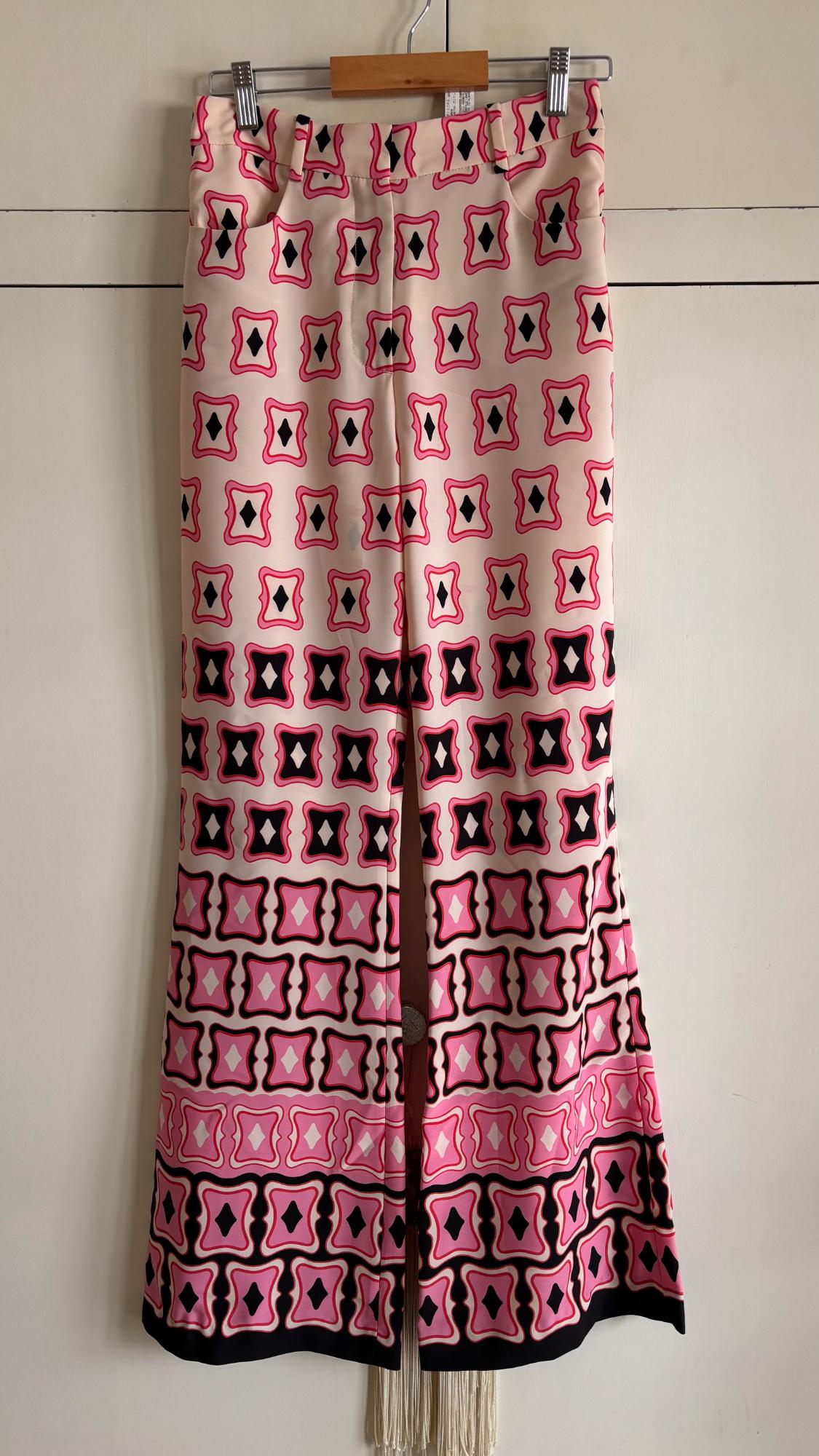 Flared trousers with a retro geometric print in pink, black, and white, hanging on a wooden hanger in front of white doors.