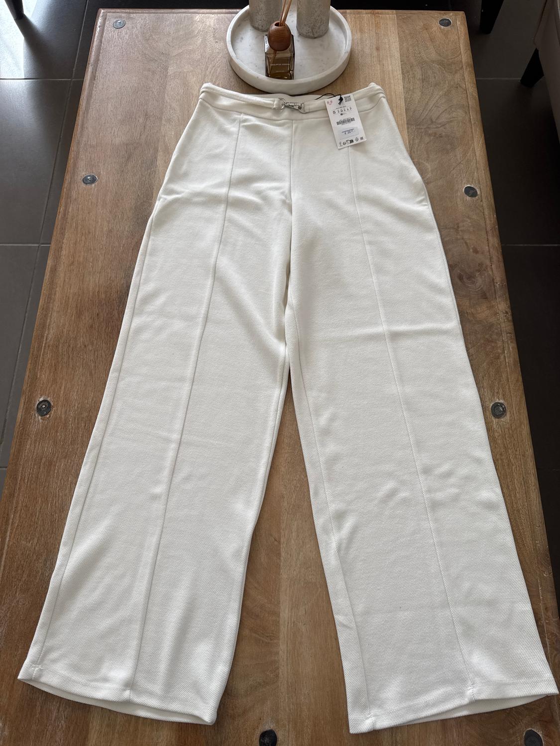 Soft white fabric trousers with a tag, placed on a wooden table. In the background, a tray with decorative objects.