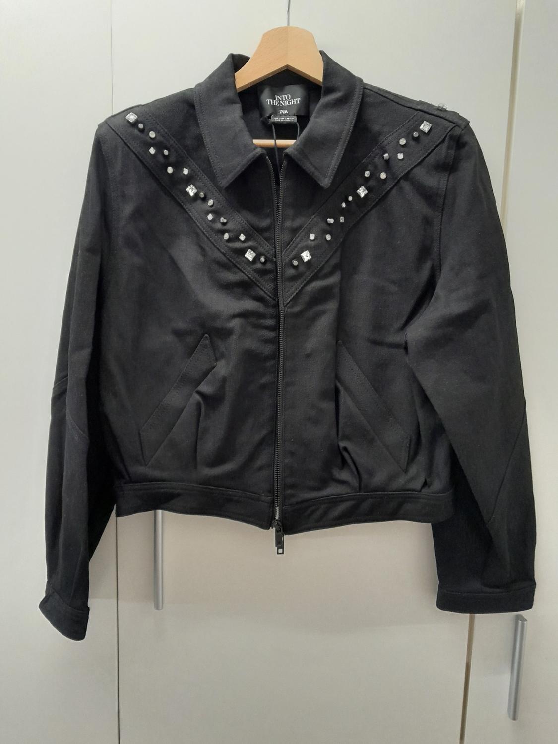 Black jacket with studs on the shoulders and chest, hanging on a wooden hanger. Visible text: "INTO THE NIGHT".